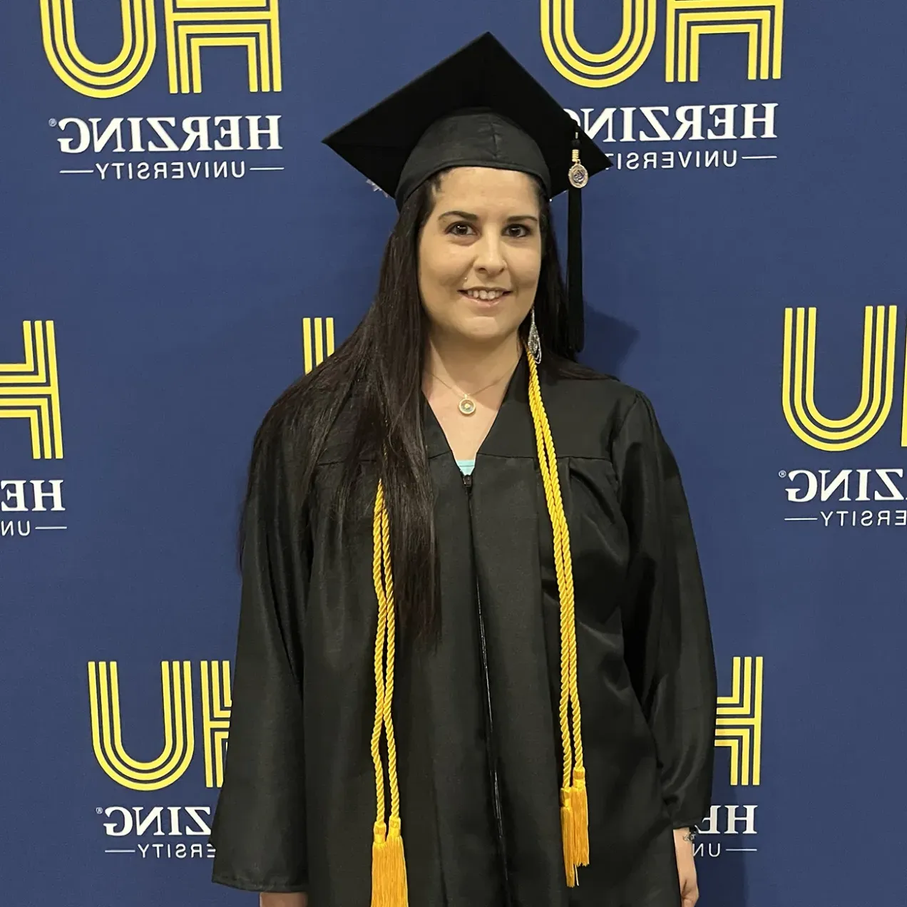 A Herzing graduate in a black graduation gown and cap stands smiling in front of a Herzing University banner, wearing two yellow honor cords around her neck.